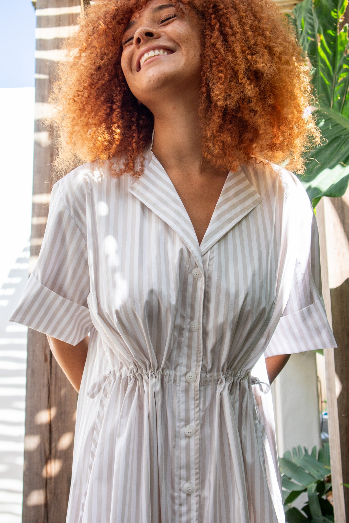 Model wearing beige stripe dress with eyes closed looking up at the sun 