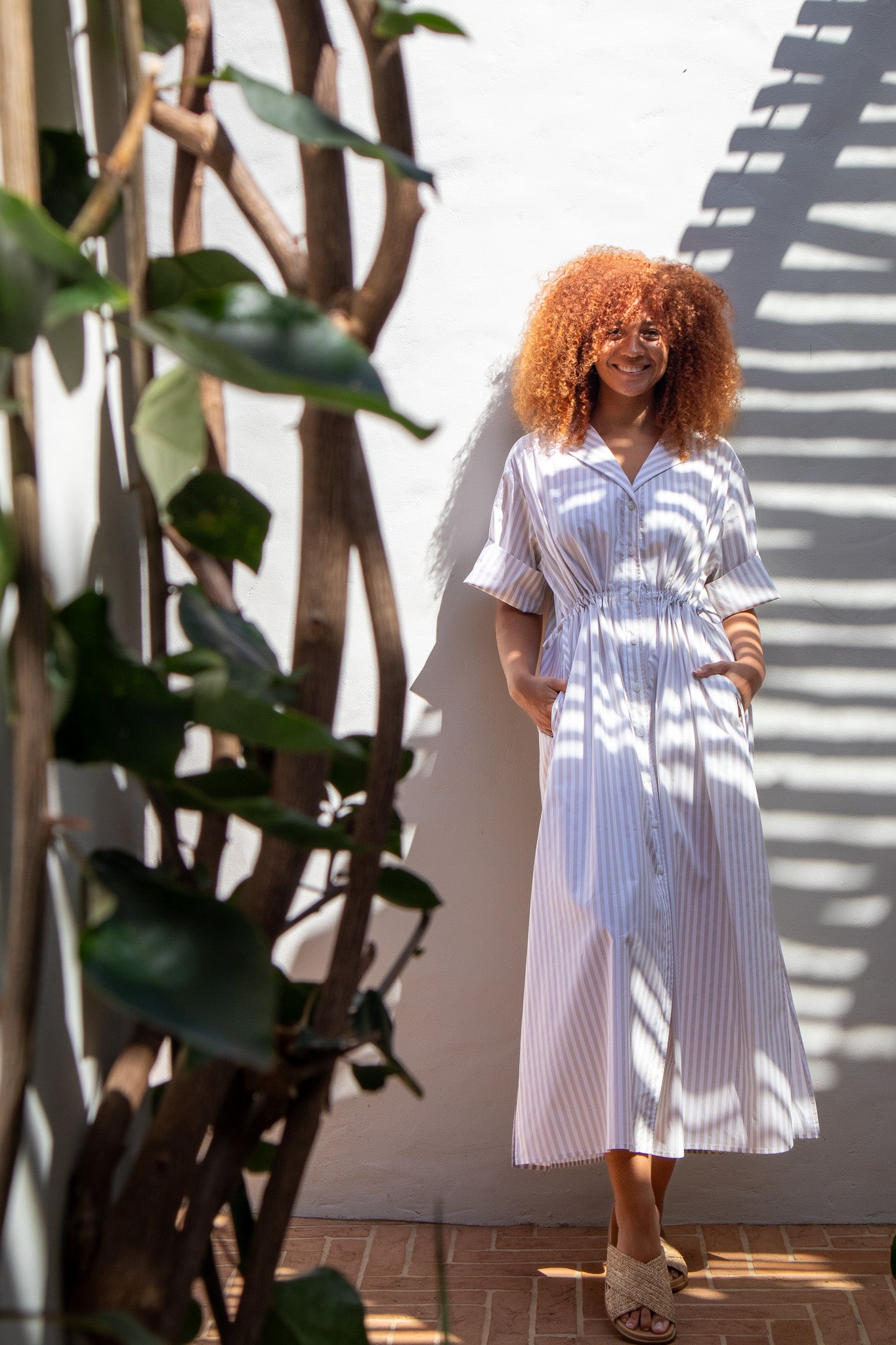 Model wearing beige stripe dress made of cotton poplin, leaning on wall with hands in pockets.