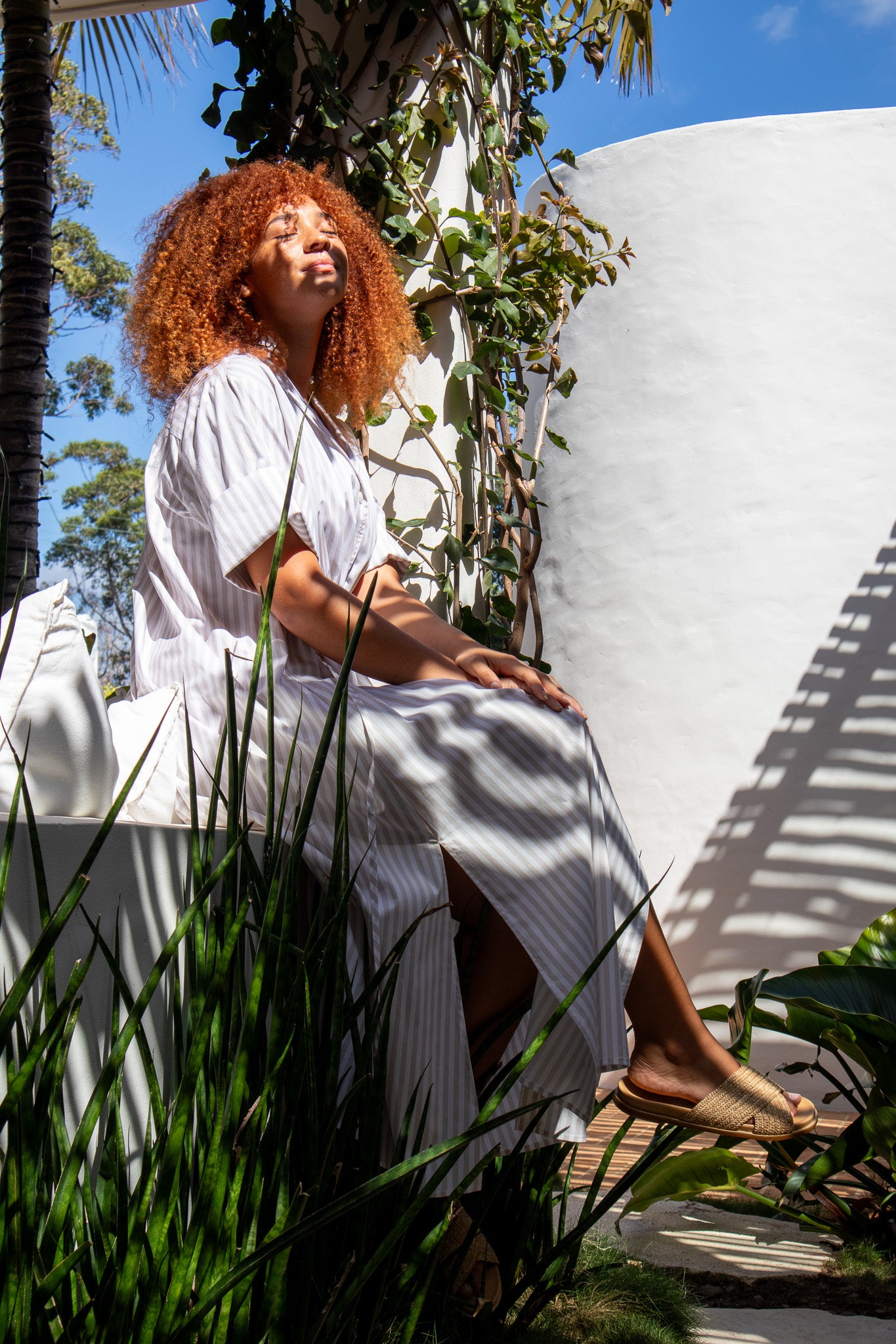 Model sitting on a wall looking up at the sun wearing a beige stripe dress from Australian clothing label, The Boy and I 